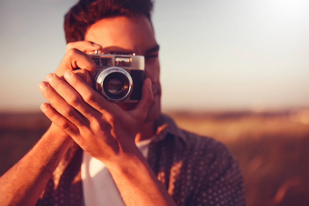 Close-up beautiful portrait of young handsome man with vintage camera, take a picture, on field background, and on sunset. Autumn, summer, mood. Photography - Foto, Bild