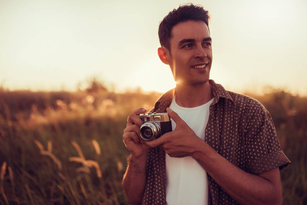 Amazing portrait of young handsome man with vintage camera look at one side, on field background, and on sunset. Autumn, summer, mood. Photography - 写真・画像