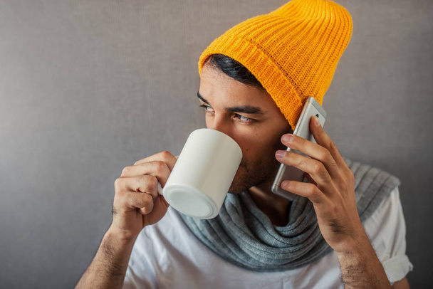 Portrait of handsome young business man sitting on gray background with a mug of coffee, tea, water. Speaking with a gadget, telephone. Wearing orange hat and gray scarf. - Fotoğraf, Görsel