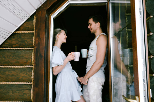Lovely couple together at home. Pretty woman and attractive man wearing stylish casual clothes standing on a balcony, drinking tea and laughing. - Fotoğraf, Görsel