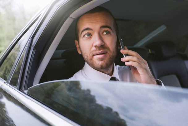 Horizontal portrait of a business man talking on mobile phone and looking out the car window in a taxi. Handosme business man using smart phone for news. Technology and business concept. - 写真・画像