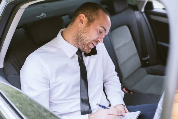 Horizontal portrait of a concentrated businessman with papers calling on smart phone and sitting on car back seat. Handsome business man wearing white shirt in black tie using mobile phone. - Φωτογραφία, εικόνα