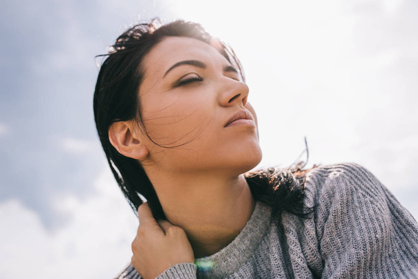 Brunette Caucasian young woman traveler with windy hair, dreaming against sunlight sky. Dreamy female with closed eyes in gray pullover. Lifestyle and people concept. Cover mood. - Foto, imagen