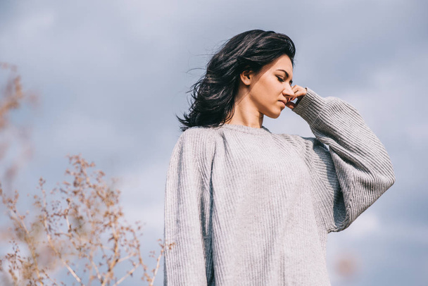 Horizontal shot of attractive brunette Caucasian young woman traveler with windy hair, posing against overcast sky. Dreamy female with closed eyes in gray pullover. Lifestyle and people concept mood. - Фото, изображение