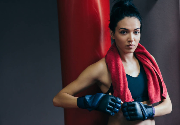 Horizontal portrait of strong attractive brunette woman with red towel on neck before workout with punch bag, wearing kickboxing gloves in the gym. Sport, fitness, lifestyle and people concept. - Foto, afbeelding