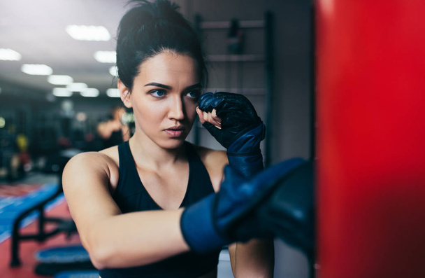 Caucasian beautiful brunette sportswoman trainer punching a red bag with kickboxing gloves in the gym. Sport, fitness, lifestyle and motivation concept. - Fotó, kép