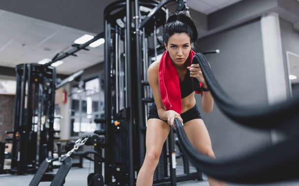 Sport, people, motivation concept. Fitness active brunette sportswoman working out in functional training gym doing crossfit exercise with battle ropes with red towel on neck, copyspace for your text - Zdjęcie, obraz