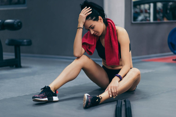Young brunette sportswoman sitting on a floor at gym after her battle rope workout and looking down. Attractive female athlete taking rest after fitness training at gym with red towel on neck. - Φωτογραφία, εικόνα