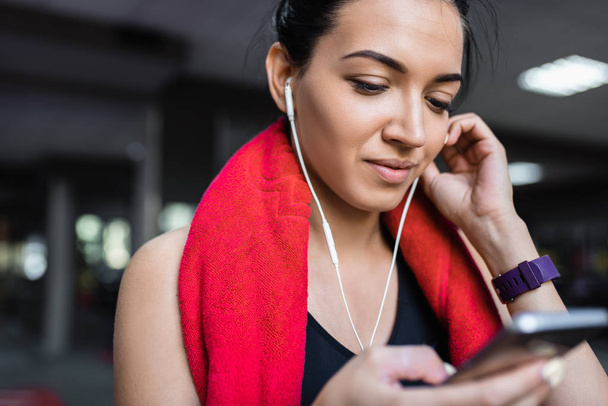 Cropped portrait of a beautiful smiling and sporty young woman listening to music from her smartphone, having a break after workout in the gym. People, technology, fitness and sport concept. - Foto, afbeelding