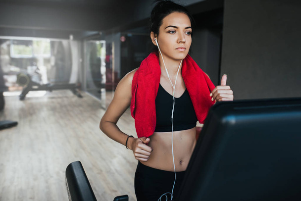 Caucasian pretty young woman athlete running on treadmill in the gym, with a red towel on neck and listening the music by her earphones. Sport, fitness, healthy life and people concept. - Photo, image