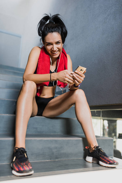 Happy and funny athletic attractive brunette woman with red towel on neck after workout in the gym sits on the stairs and listening the music on earphones from smartphone. Sport concept. - Photo, Image