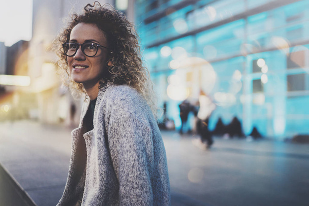 Smiling woman in stylish clothing wearing eye glasses outside in the european night city. Bokeh and flares effect on blurred background. - Photo, image