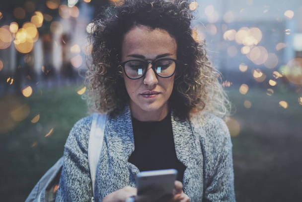 Female hipster wearing backpack and glasses searching information in mobile network by smart phone, during walk in european city at the night. Bokeh and flares effects on blurred background. - Фото, изображение