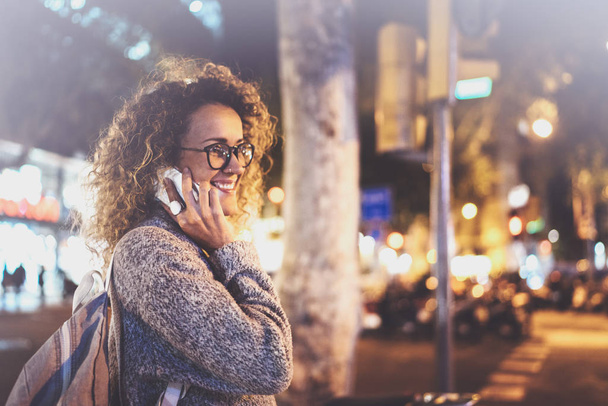 Smiling pretty woman making conversation call with her friends via cell telephone while standing at night on the street. Bokeh and flares effect on blurred background. - Fotoğraf, Görsel