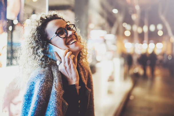 Smiling pretty woman making conversation call with her friends via cell telephone while standing at night on the street. Bokeh and flares effect on blurred background. - Fotografie, Obrázek