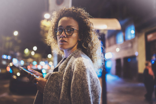 Beautiful girl hipster wearing glasses holding smart phone hands, during night walking in european vocation. Bokeh and flares effects on blurred background. - Fotó, kép