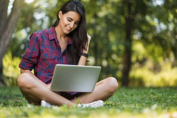 Attractive shot of young brunette Caucasian female with a smile freelancer dressed casual, sitting on the lawn in front of notebook, working on laptop computer, shopping, reading articles, studying. - Foto, Imagem