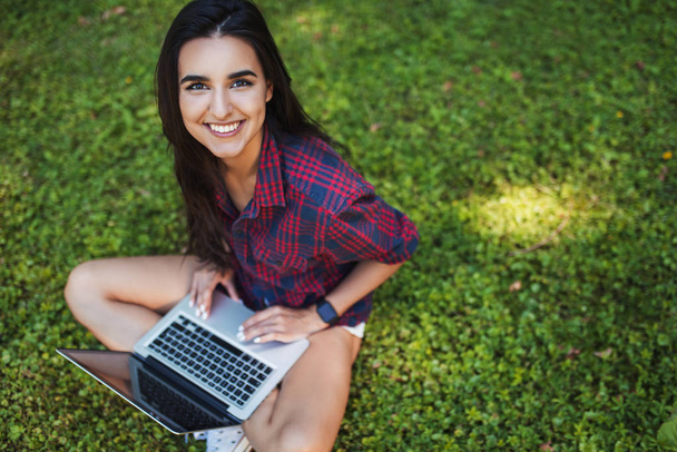 Top view of young Caucasian brunette woman smiling and wearing plaid shirt, sitting on grass in front of laptop pc, working on her notebook computer with wireless communication, shopping and studying - Zdjęcie, obraz