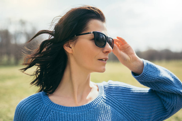 Portrait of a young brunette happy woman in sunglasses at the park. Caucasian happy attractive female outdoors in the spring on sunshine day, looking away. Pretty girl with windy hair, freedom view. - Photo, Image