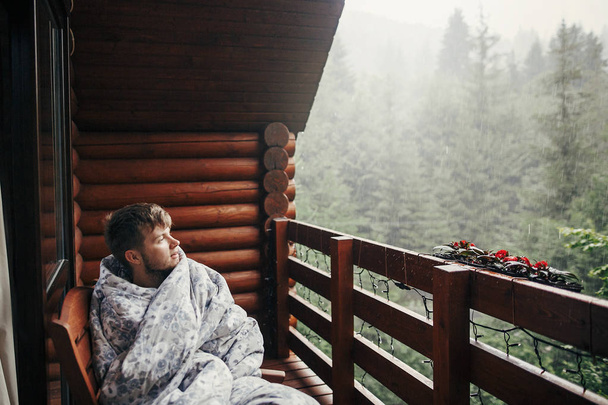 stijlvolle bebaarde man ontspannen op houten veranda onder bos in Regenachtig Bergen. de kerel van de Hipster rusten in deken, zittend in huisje, kijken naar regen. rustige vreedzame moment - Foto, afbeelding