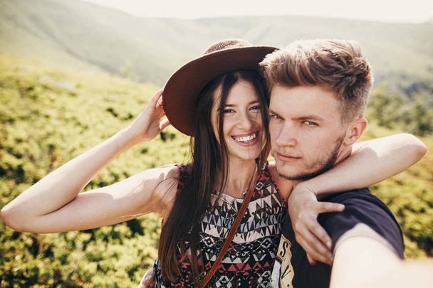 stylish hipster couple taking selfie and smiling on top of mountains in sunny summer day. beautiful man and happy woman in hat hugging and embracing on mountain. travel together - 写真・画像