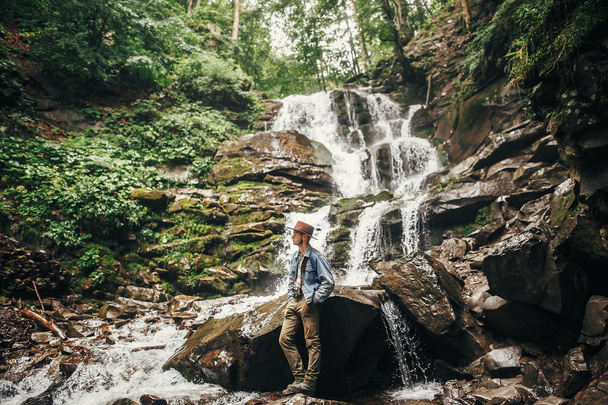 stylish hipster man in hat with photo camera, standing at waterfall in forest in mountains. traveler guy exploring woods. travel and wanderlust concept. atmospheric moment - Photo, Image