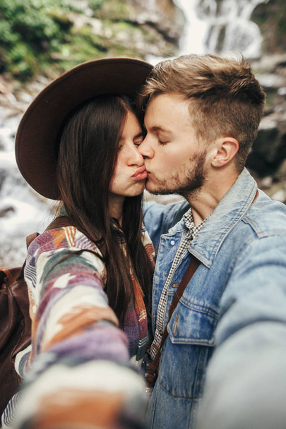 stylish hipster couple taking selfie and kissing at waterfall in forest in mountains. handsome man and happy woman in hat embracing on honeymoon. travel together. summer vacation - Foto, Bild