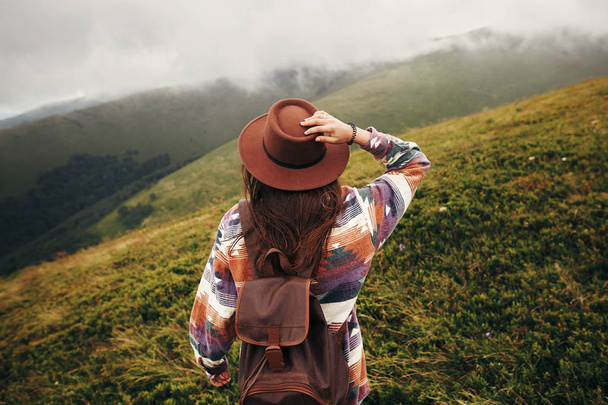 stijlvolle reiziger hipster meisje houden hoed, met rugzak en winderige haren, wandelen in de bergen in de wolken. ruimte voor tekst. geweldig sfeervolle moment. reizen en wanderlust concept - Foto, afbeelding