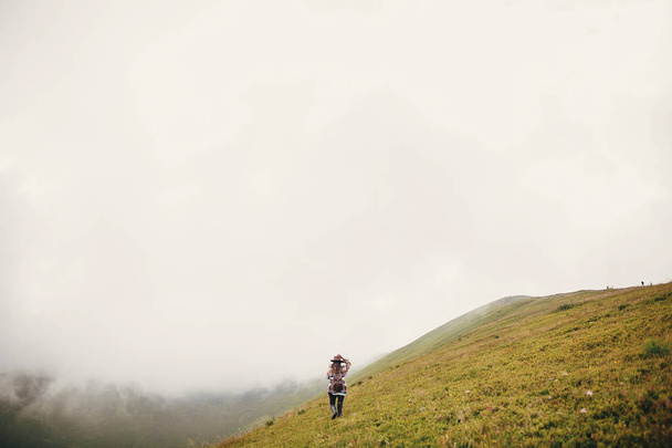Stilvolles Reiselädchen mit Hut, Rucksack und windigen Haaren, das in den Wolken der Berge wandert. Sommerferien. Reise- und Fernweh-Konzept. Raum für Text. Stimmungsvoller Moment - Foto, Bild
