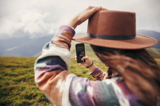 dumdum dziewczyna w kapeluszu wietrznie włosy trzymając telefon na szczycie góry sunny. letnie wakacje. koncepcja podróży i wanderlust. miejsca na tekst. Widok z tyłu. niesamowity moment atmosferyczne - Zdjęcie, obraz