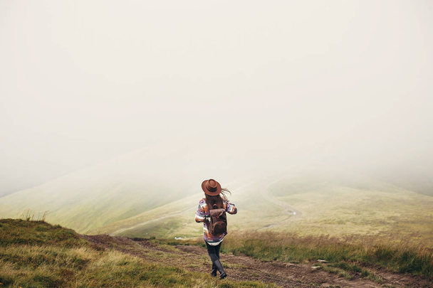 reizen en wanderlust concept. stijlvolle reiziger meisje houden hoed, met rugzak en winderige haren, wandelen in de bergen in de wolken.  - Foto, afbeelding