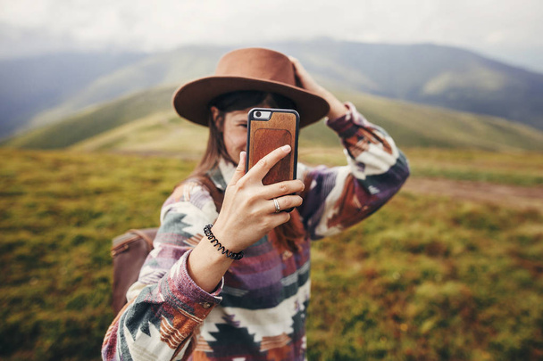 menina viajante elegante em chapéu com cabelo ventoso segurando telefone e fazendo selfie no topo das montanhas. viagem e conceito de luxúria
.  - Foto, Imagem