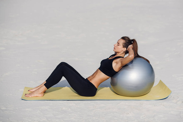 Sport and healthy lifestyle concept. Beautiful fitness girl doing stomach exercises with fit ball on the beach. Young woman doing pilates exercises. - Fotografie, Obrázek