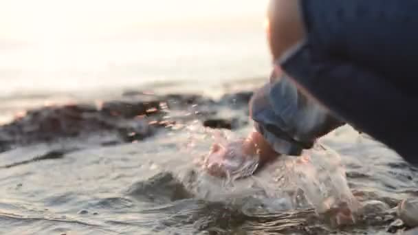 Uma menina salpicando água do mar pela costa do mar. Close-up. Pôr do sol no mar. Movimento lento
 - Filmagem, Vídeo
