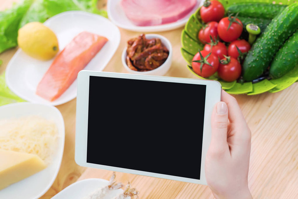 Close-up top view of female hands holding a tablet computer with a blank display surrounded by healthy food on a home kitchen table. Workpiece to insert the design of the mobile application into the - Photo, image