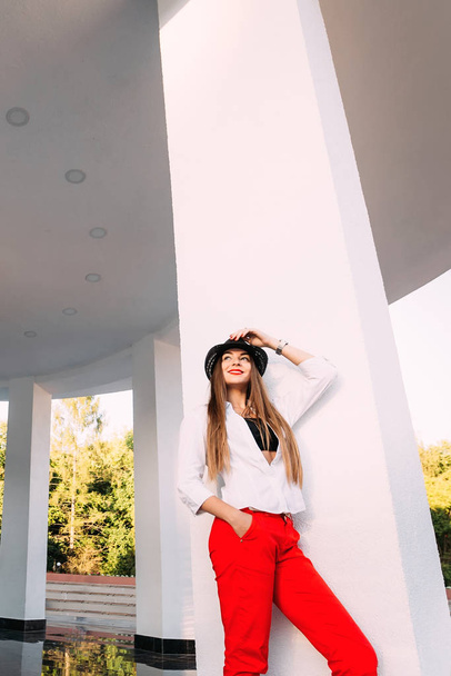дівчина стоїть біля колони будівлі в парку і тримає руки на чорному капелюсі, а інша рука в кишені
 - Фото, зображення