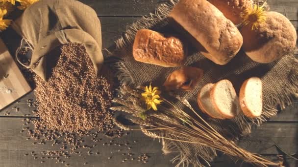 Still life with bread, wheat, flour and flowers.Men's hands take a handful of wheat grains from a sack and pour them back into the bag.Slow motion. Top view. Horizontal ( from right to left ) pan.  - Footage, Video