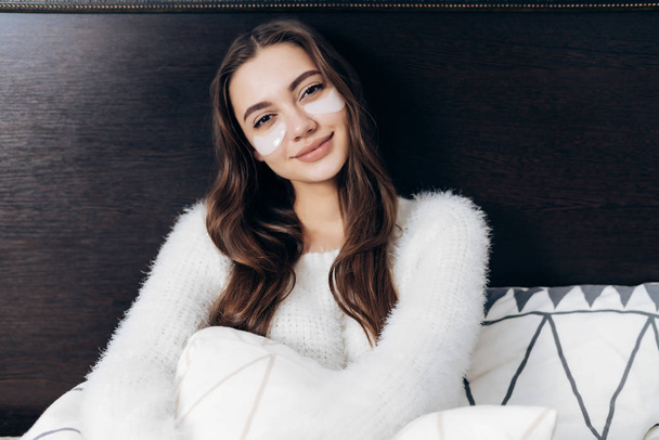hermosa chica de pelo largo en pijama blanco se sienta en su cama temprano en la mañana, bajo los ojos manchas blancas
 - Foto, imagen