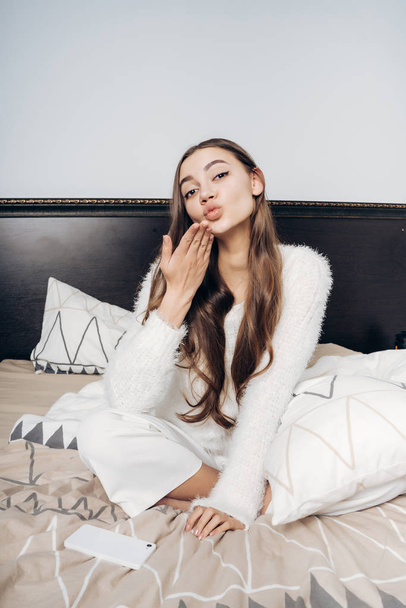 cute young long-haired girl in white pajamas sits on the bed and sends an air kiss - Фото, изображение