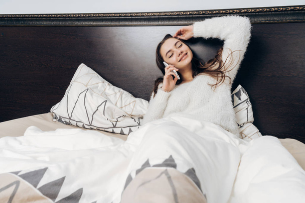 happy young girl in pajamas lying in bed, resting after working day, talking on phone and smiling - Valokuva, kuva