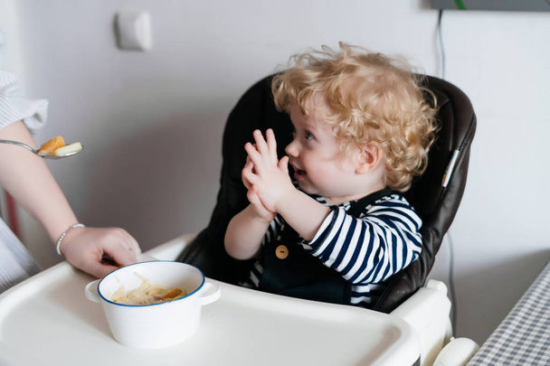 a small curly child is having breakfast with his mother, he is happy, useful porridge - Фото, изображение