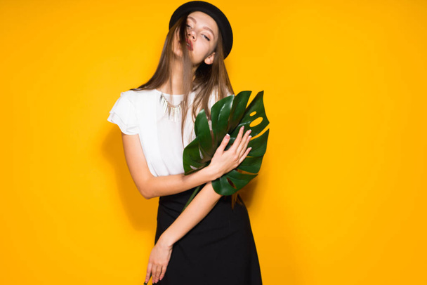 attractive long-haired girl model in black hat holds a green leaf, posing on orange background - 写真・画像