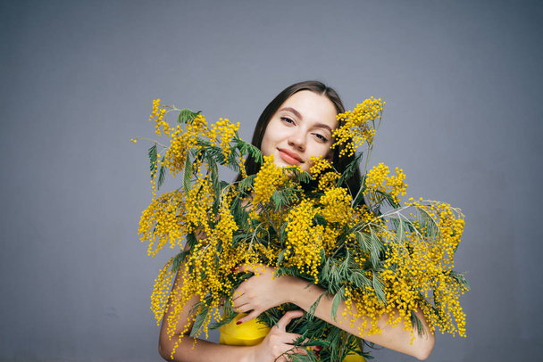 happy young girl in a yellow dress holds a big bouquet of a fragrant yellow mimosa, enjoys spring and warmth - Фото, изображение