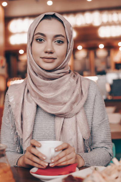 beautiful Arab girl with headscarf sitting in cozy cafe, drinking coffee, smiling and looking at camera - Photo, image