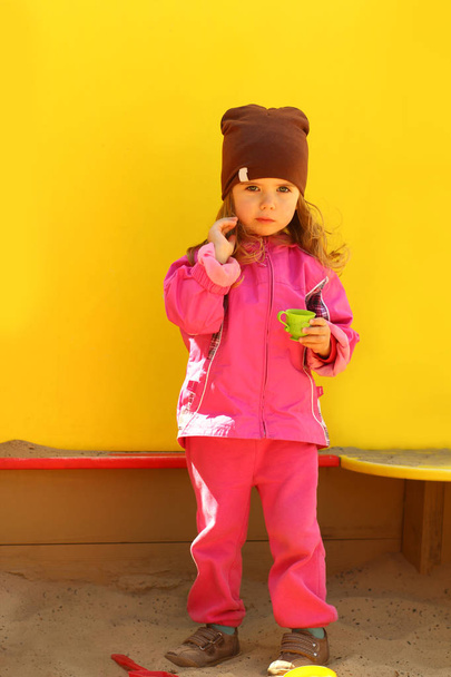Güneşli parlak sarı arka plana karşı kum kutusunda "kahve" içme yürümeye başlayan çocuk kız - Fotoğraf, Görsel