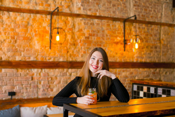 Ικανοποιημένοι κορίτσι που κάθεται κοντά στο ξύλινο τραπέζι με φλιτζάνι λεμονάδα σε καφέ και ξεκούραση. - Φωτογραφία, εικόνα