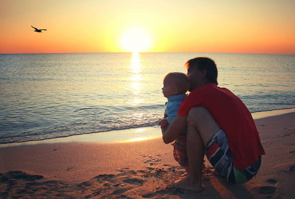 Vader en zijn zoontje van de baby, zittend op zonsondergang strand. Familie vakantie aan de zee. - Foto, afbeelding