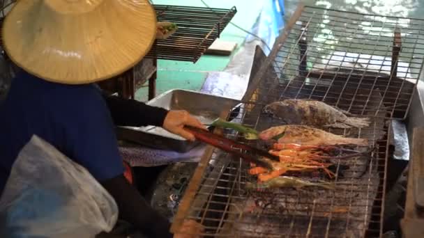 Een Thaise vrouw in traditionele doek koken grote garnalen op een Barbecue-brand in de boot op de drijvende markt, Bangkok. - Video