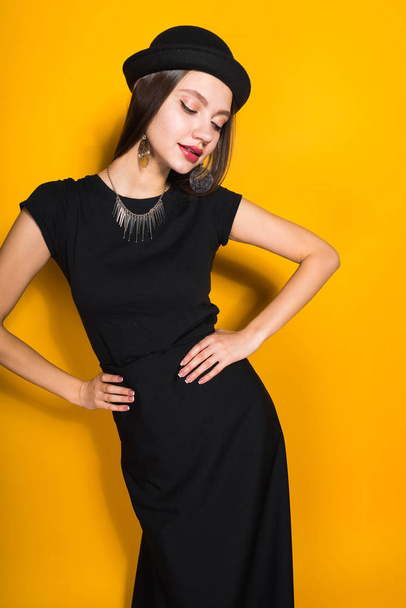 confident attractive girl in long black dress and fashionable hat posing on yellow background - Fotografie, Obrázek