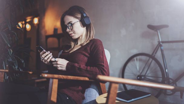 Young attractive girl wearing eye glasses and casual clothes listening digital music in headphones on smartphone at modern place. - 写真・画像